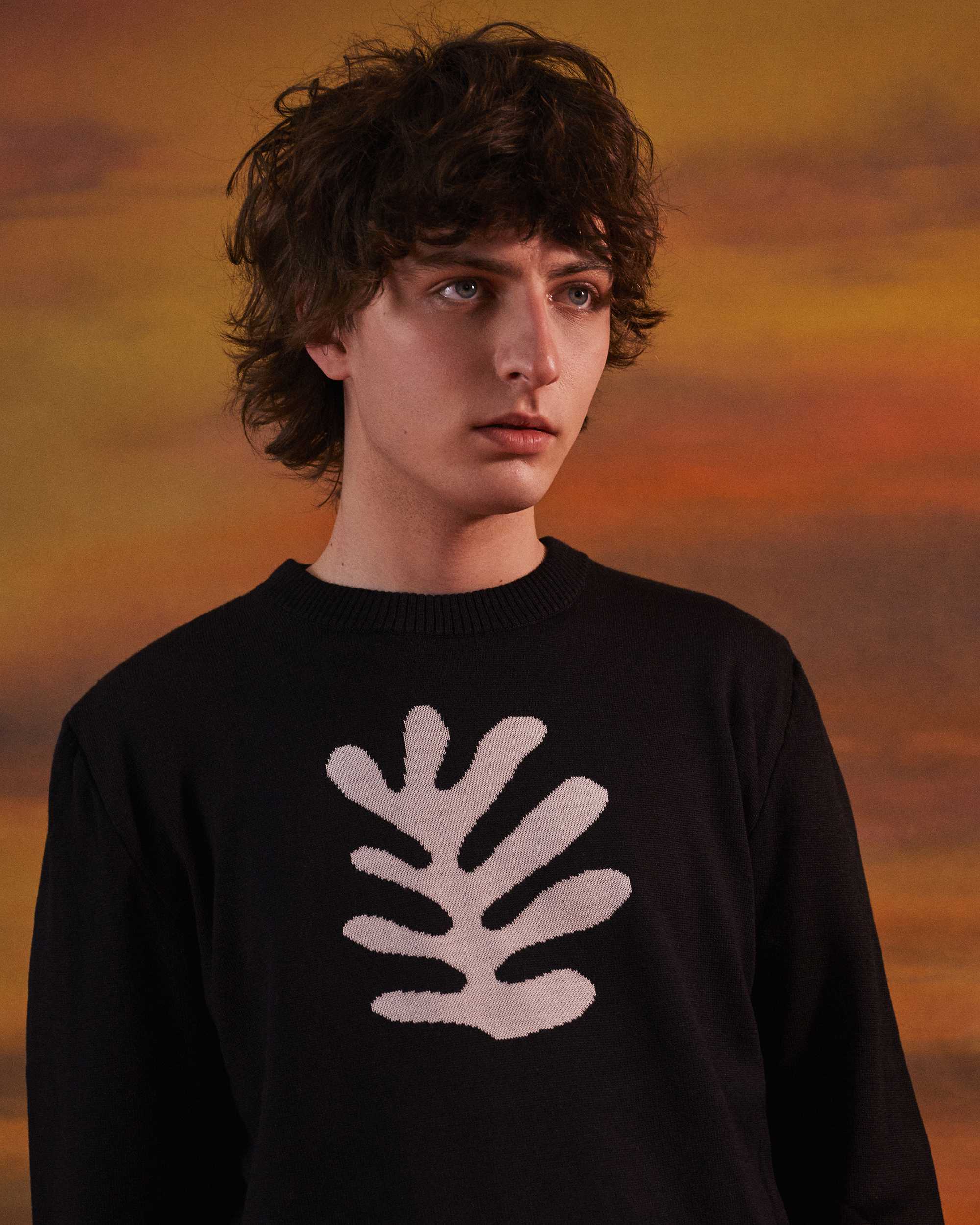 Man posing infront behind a sunset in a black knitted jumper with a white organic shape on