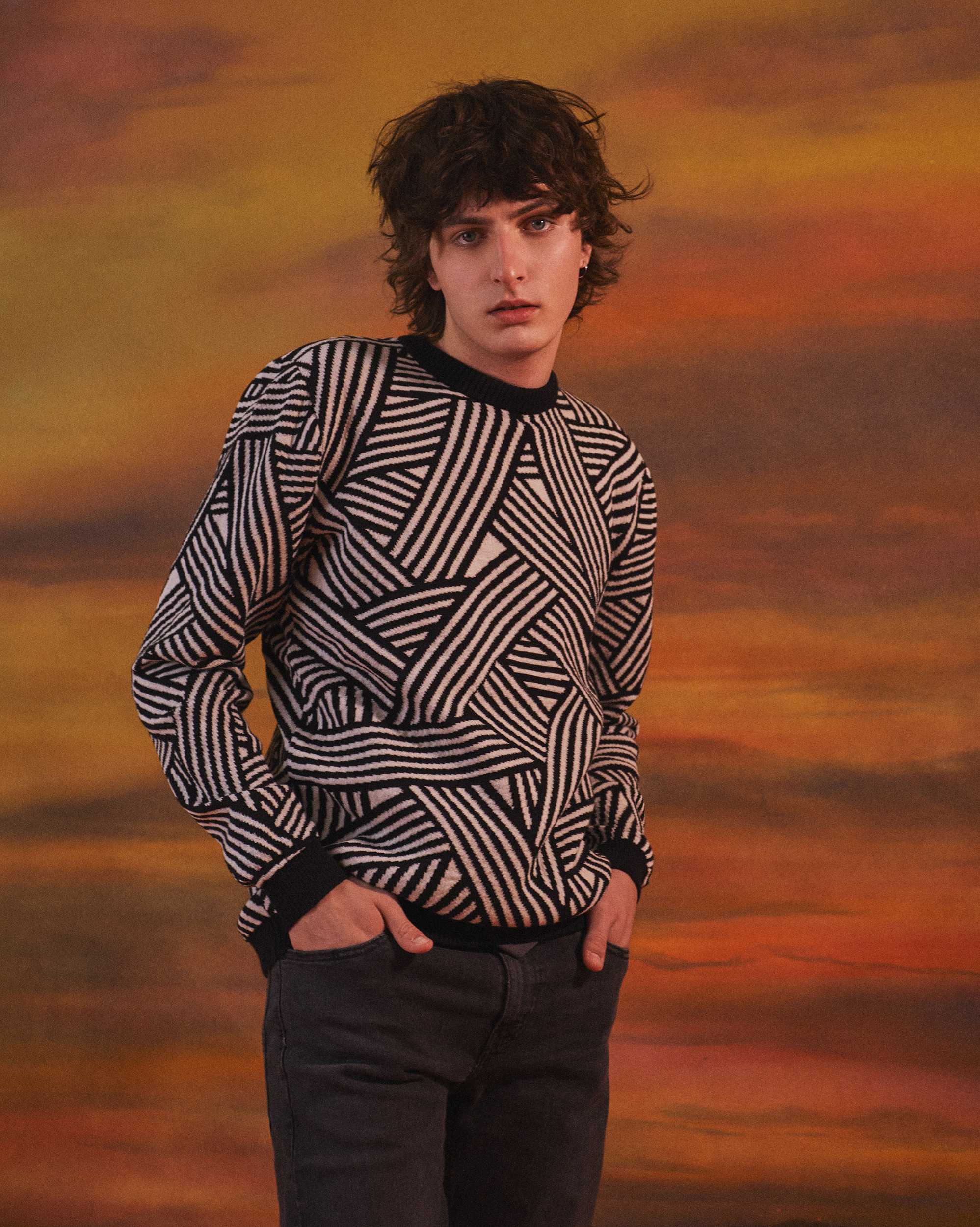 Man standing infront of sunset in a stripy black and white knitted jumper and black jeans 