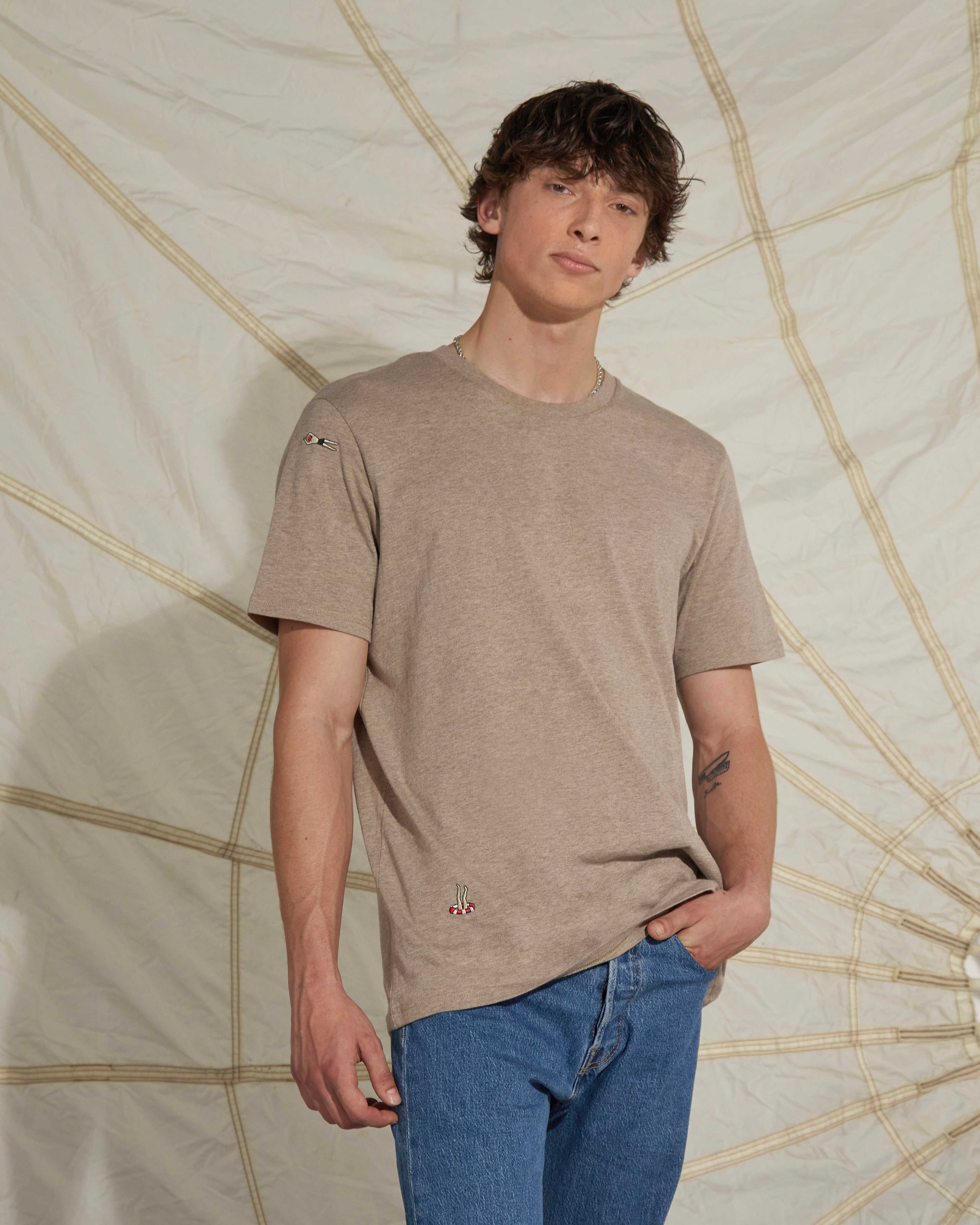Man wearing a beige tshirt with swimmers embroideries and blue levis jeans posing infront of a kite
