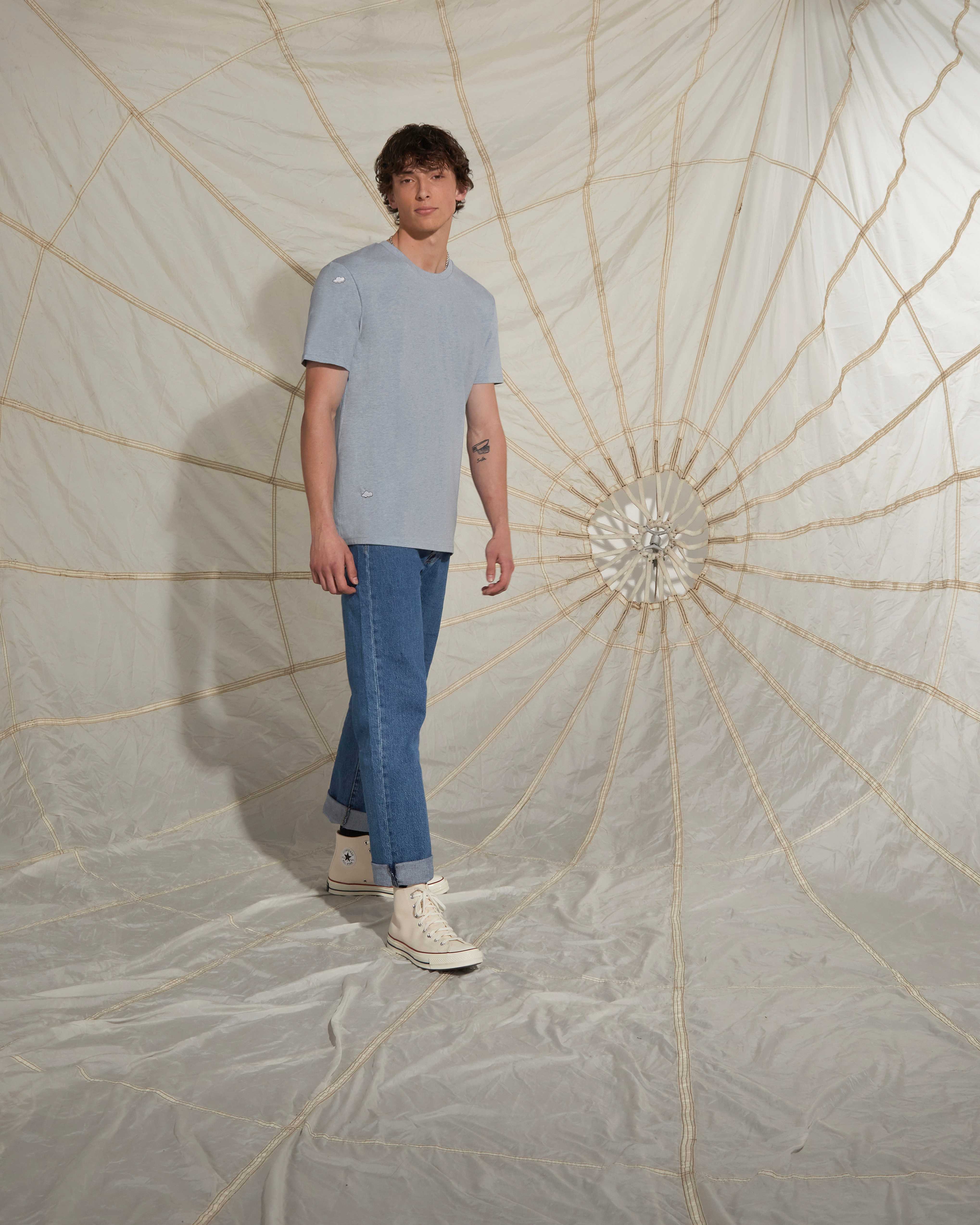 A man wearing a blue ingmarson top with clounds embroidered matched with blue levis jeans and cream converse shoes posing infront of a parachute. 