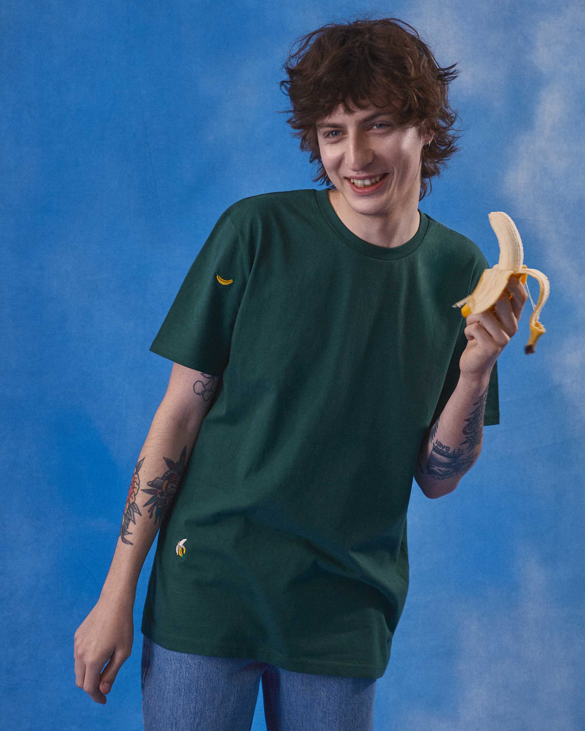 Man standing infront of a blue sky eating a banana in a green t-shirt with banana emroideries on
